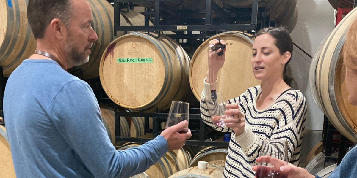 Guest holding up a wine glass in a barrel room filled with a wine thief by a winemaker